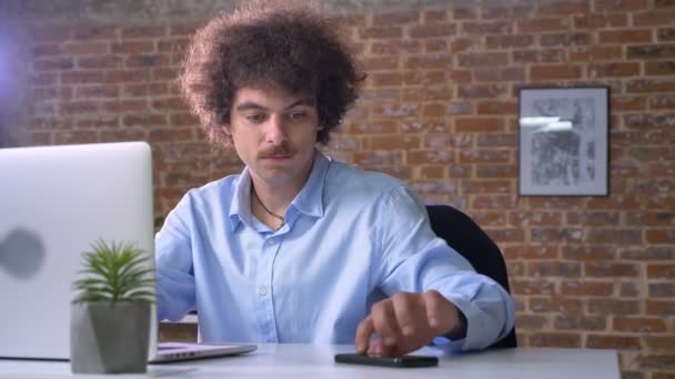Deprimido nerd chefe com cabelo encaracolado tendo problemas com o negócio, trabalhando no laptop e tomando telefone, moderno escritório fundo — Vídeo de Stock