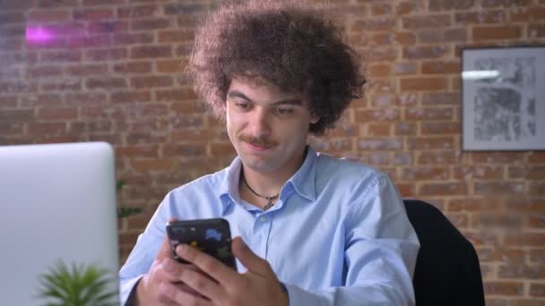 Αστεία nerdy επιχειρηματίας με όγκο σγουρά μαλλιά κατάδειξης τηλέφωνο με chroma κλειδί σε κάμερα, κάθεται και εργάζεται στο σύγχρονο γραφείο — Αρχείο Βίντεο