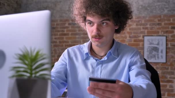 Empresário nerd engraçado com cabelo encaracolado compras através da internet no laptop e pagar com cartão de crédito, sentado no escritório — Vídeo de Stock