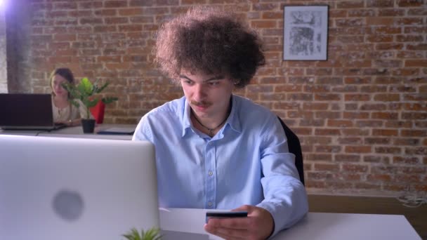 Щасливий смішний офісний працівник купує через інтернет на ноутбуці і використовує свою кредитну картку, веселий чоловік в сучасному будинку, в приміщенні — стокове відео