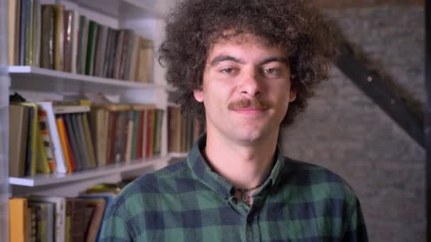 Rolig nerdy människa med lockigt hår och mustasch står i biblioteket och titta på kameran, glad och positiv — Stockvideo