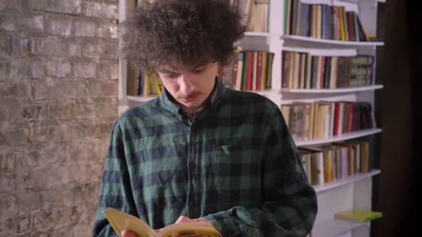 Sprytny student nerdy z kręconymi włosami czytania książki w bibliotece i stałego, regały w tle — Wideo stockowe