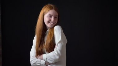 Siyah arka plan üzerine izole kamera, gülümseyen beyaz gömlekli uzun kızıl saçlı utangaç büyüleyici kadın