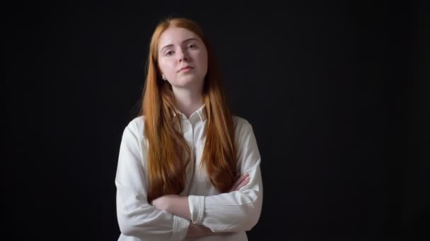 Serious young woman with long ginger hair looking at camera with crossed hands, isolated on black background — Stock Video