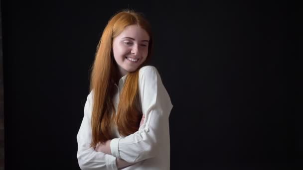 Tímida mujer encantadora con el pelo largo de jengibre en camisa blanca sonriendo a la cámara, aislado sobre fondo negro — Vídeos de Stock