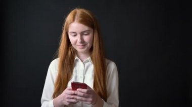 Büyüleyici zencefilli kadın telefon ve dokunaklı onun saç, izole üzerinde siyah arka plan üzerinde beyaz gömlek manifatura