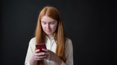 Telefona yazma ve siyah arka plan üzerine izole kamera, gülümseyen uzun kızıl saçlı Sevimli genç kadın