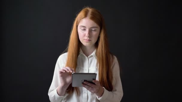 Genç Genç kız beyaz gömlekli tablete yazarak zencefil ve ayakta kameraya seyir siyah arka plan üzerine izole — Stok video
