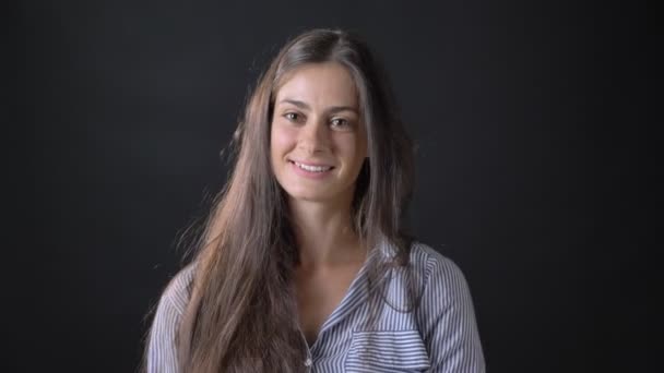 Encantadora joven con camisa a rayas sonriendo a la cámara y tocándose el pelo, de pie aislada sobre fondo negro — Vídeos de Stock