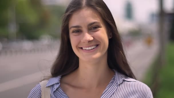 Hermosa joven feliz mujer sonriendo a la cámara y tocando el pelo, de pie en la calle cerca de la carretera — Vídeos de Stock