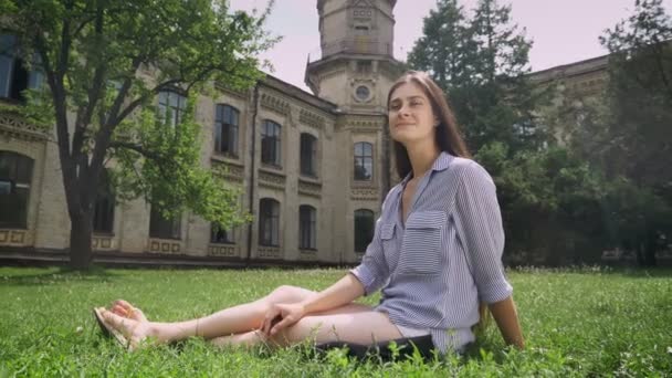 Encantador jovem estudante universitário com cabelos longos sentado na grama no parque perto da universidade e sorrindo, feliz e alegre — Vídeo de Stock