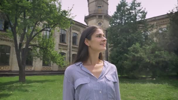 Alegre joven bonita mujer en camisa girando la cabeza y mirando a la cámara, sonriendo, de pie en el parque cerca de la universidad — Vídeos de Stock