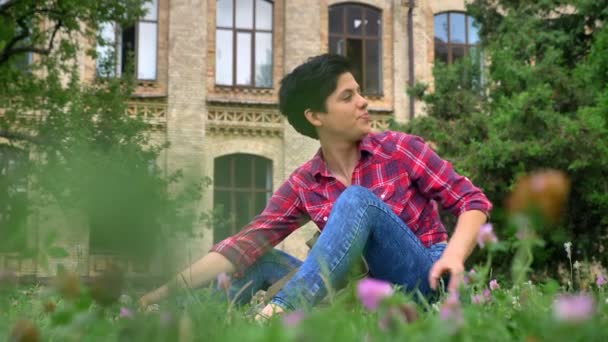 Okouzlující mladý vysokoškolský student s černými krátkými vlasy sedí na trávě v parku nedaleko univerzity, šťastný a veselý — Stock video