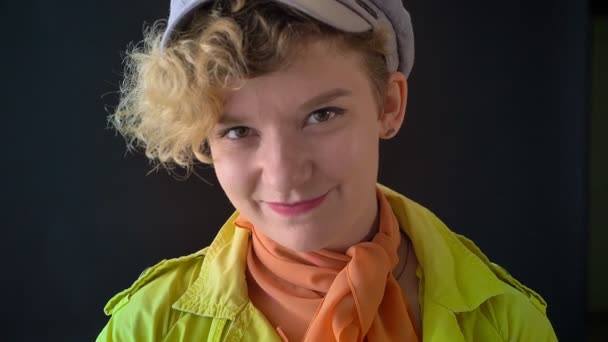 Giovane donna felice con i capelli biondi ricci in cappello guardando la fotocamera e sorridente, isolato su sfondo nero studio — Video Stock