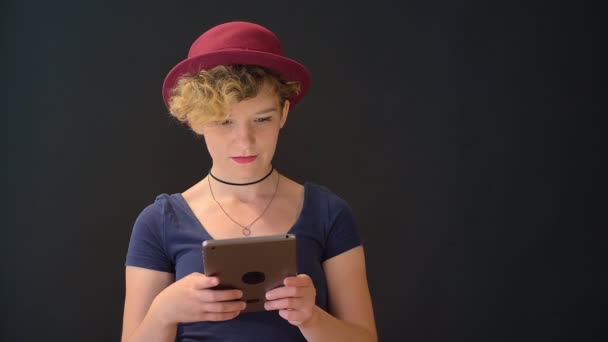 Ernstige jonge mooie vrouw in rode hoed met krullend haar bedrijf van tafel en kijken naar camera, geïsoleerd op zwarte achtergrond — Stockvideo