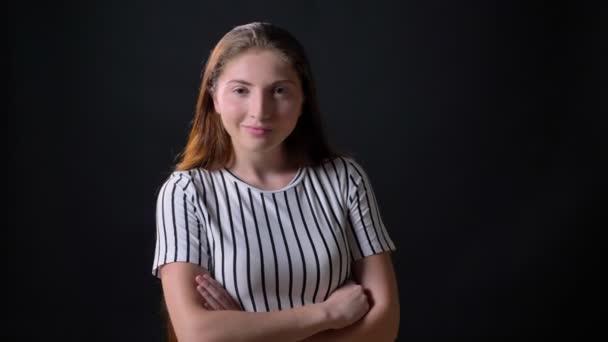 Självsäker ung kvinna i randig skjorta ler mot kameran och stående med korsade händer isolerad på svart bakgrund — Stockvideo
