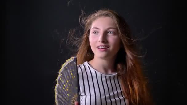 Joven mujer feliz con el pelo largo de jengibre ventilarse con ventilador, sonriendo, expresión sorprendida, aislado sobre fondo negro — Vídeos de Stock