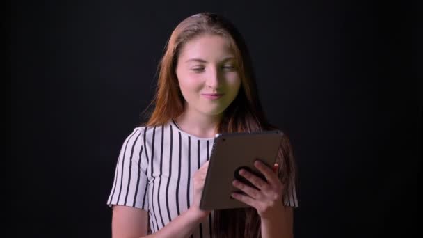 Красива імбирна жінка посміхається і тримає планшет з хромакією, стоїть ізольовано на чорному студійному фоні — стокове відео