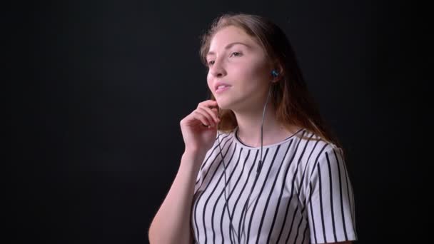 Mujer encantadora feliz soñadora escuchando música a través de auriculares y sonriendo, de pie aislado en el fondo del estudio negro — Vídeo de stock