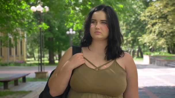 Συγχέεται νεαρή γυναίκα με το υπερβολικό βάρος γύρω και στέκεται στον δρόμο, στο πάρκο κατά τη διάρκεια της ηλιόλουστη μέρα — Αρχείο Βίντεο