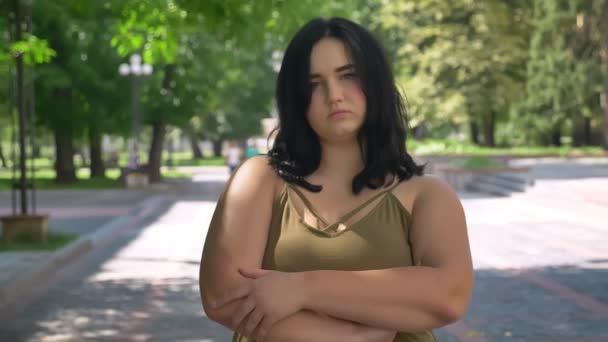 Graves deprimido joven bonita mujer con obesidad mirando a la cámara, de pie en la calle en el parque durante el día soleado — Vídeos de Stock