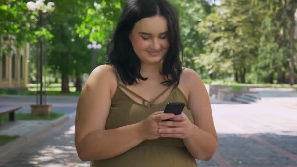 Piękna Brunetka kobieta otyłych wpisując na telefon i stojąc na ulicy, w parku, w pobliżu Uniwersytetu, słoneczny dzień na zewnątrz — Wideo stockowe