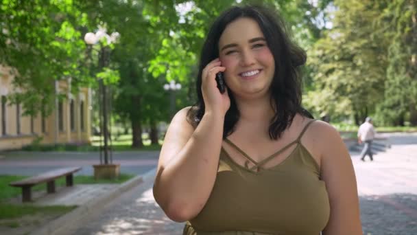 Красивая молодая толстая женщина разговаривает по мобильному телефону и улыбается, стоит на улице в парке, счастливая и веселая — стоковое видео