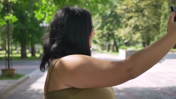 Mulher obesa feliz bonita jovem ter vídeo chat através do telefone e sorrindo, de pé na rua no parque — Vídeo de Stock