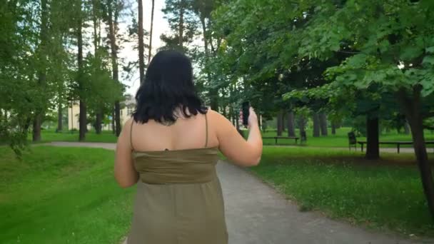 Вид ззаду красива молода жінка, яка бере селфі і ходить в парку з деревами і зеленою травою, чарівна леді в одязі — стокове відео