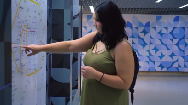Mulher obesa bonita em pé no metrô subterrâneo moderno e olhando para o mapa, encantador turista feminino com mochila — Vídeo de Stock