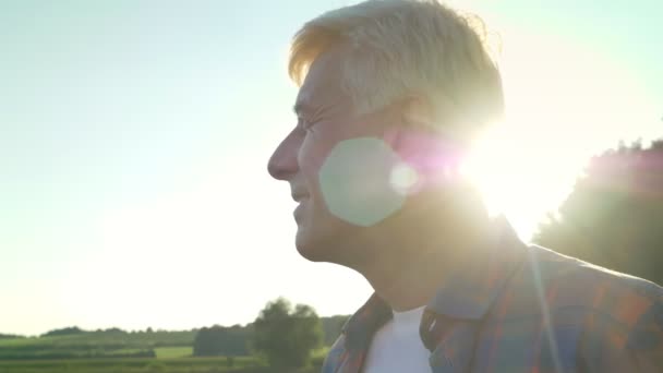 Feliz velho agricultor masculino olhando para a frente em seu campo e sorrindo, bela natureza durante o pôr do sol no fundo, chama lente — Vídeo de Stock