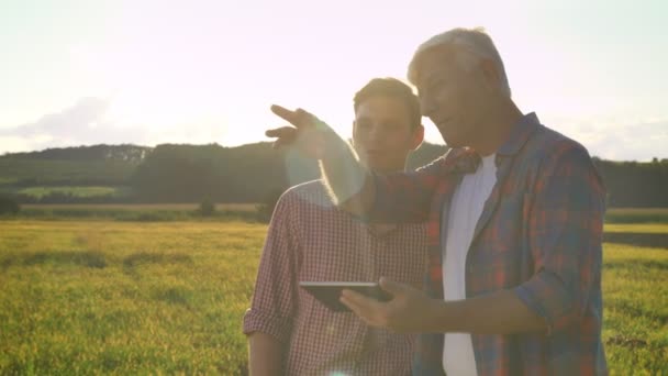 Velho agricultor digitando em tablet e mostrando ao seu herdeiro campo de trigo, ensinando filho sobre agricultura, bela vista durante o pôr do sol — Vídeo de Stock