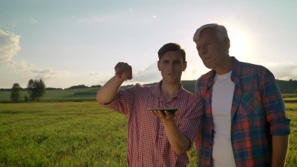 Filho adulto segurando tablet e discutindo novo projeto com seu pai velho, de pé no campo de trigo durante o belo pôr do sol no fundo — Vídeo de Stock