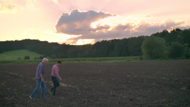 Взрослый сын гуляет со своим старым отцом на почвенном поле, красивый вид на обрабатываемые земли во время заката на заднем плане — стоковое видео