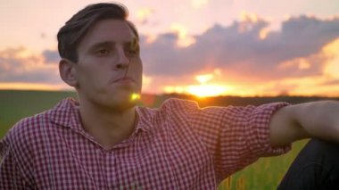 Dört gözle, arka planda günbatımı güzel manzaralı buğday alanında oturup ve saman, Çiğneme yakışıklı genç adam