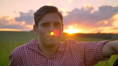 Gülümseyen ve kamera, arka planda günbatımı güzel manzaralı bakarak buğday alanında, oturup kendine güvenen yakışıklı genç