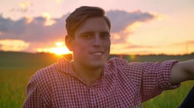 Güzel genç gülümseyen ve buğday alanında oturup, doğa günbatımı arka planda ile inanılmaz fotoğraf makinesi, bakıyorsun adam