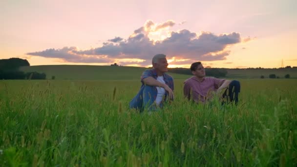 Glücklicher alter Vater und sein erwachsener Sohn sitzen auf einem Weizen- oder Roggenfeld und lächeln, schöner Sonnenuntergang im Hintergrund — Stockvideo