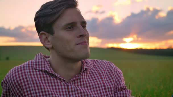 Retrato de joven hombre guapo mirando hacia adelante y sonriente, hermoso paisaje con puesta de sol en el fondo — Vídeos de Stock