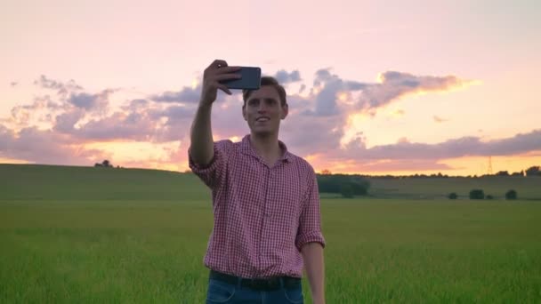 Schöner junger Mann macht Selfie und steht auf einem Weizen- oder Roggenfeld, schöner rosa Sonnenuntergang im Hintergrund — Stockvideo