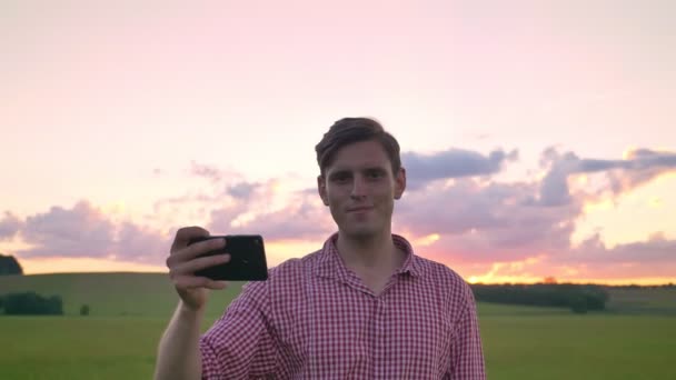 Kjekk ung mann som tar selfie og ser på kamera, står på hveteåkeren, vakker rosa himmel i bakgrunnen – stockvideo
