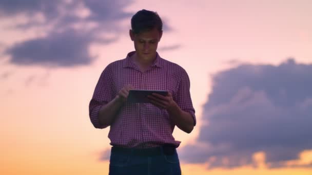 Guapo joven confiado en la camisa escribiendo en la tableta y mirando hacia adelante, aislado en el cielo rosa con fondo de la puesta del sol — Vídeos de Stock