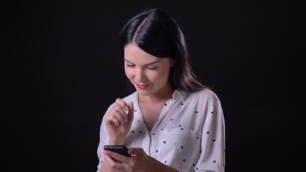 Чарівна молода брюнетка, що друкує по телефону, стоїть ізольовано на чорному тлі, посміхається на камеру — стокове відео