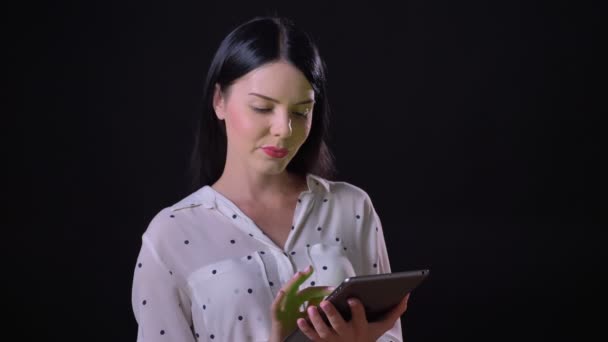 Charmante jonge brunette vrouw weergegeven: tablet met chromakey en lacht op camera, staande geïsoleerd op zwarte achtergrond — Stockvideo