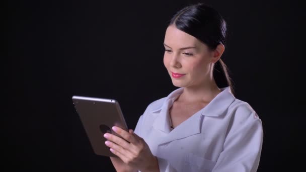 Belle jeune femme médecin en manteau blanc tapant sur tablette et debout isolé sur fond de studio noir — Video