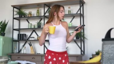 Yürekleri komik pantolon giyen kahve içme, telefonla yazarak ve modern mutfakta dans güzel kızıl kadın