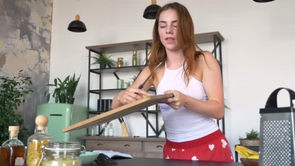 Aantrekkelijke jonge vrouw met gember haar opheffing snijplank, eten van banaan en neerzetten van een segment, moderne keuken op achtergrond — Stockvideo