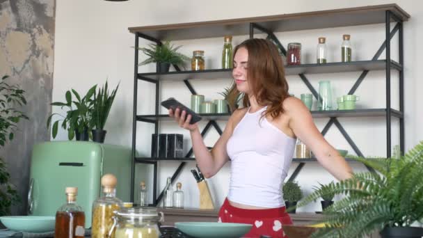 Beautiful ginger woman typing phone and dancing in modern kitchen, perfect female body, female reading recipe from smartphone — Stock Video