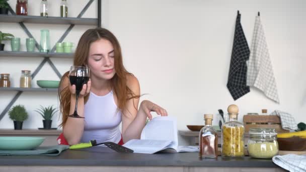 Gyönyörű gyömbér nő kulináris könyvet olvas, a gazdaság pohár bort és a modern konyha, boldog és vidám tánc — Stock videók