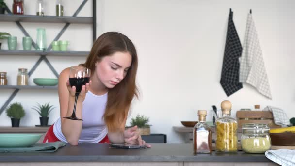 Gorgeous ginger woman typing on phone and holding glass of wine, standing in kitchen with elbows on table, dancing and relaxing — Stock Video